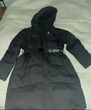 Svart lång pufferjacka Calvin Klein - Svart lång pufferjacka från Calvin Klein Jeans med huva och bälte med logga. Jackan har quiltad design, dragkedja och långa ärmar. Materialet är polyester och syntet, perfekt för kalla dagar. Snygg CK-detalj på bröstet och insidan.