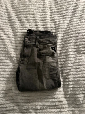 Replay grå jeans slim fit - Snygga grå jeans från Replay med slim fit och klassisk femficksdesign. Byxorna har svarta detaljer på bakfickan och Replay-logga på linningen. Materialet är mjukt och stretchigt jeans, perfekt för dig som gillar en smal siluett och stilren look.