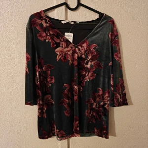Blommig mörkgrön sammetstopp KappAhl - Säljer en snygg mörkgrön topp från KappAhl i mjuk sammet med stora blommor i vinröda och rosa toner. Toppen har v-ringad hals och trekvartsärm, perfekt för dig som gillar lite lyxig känsla och coolt mönster.