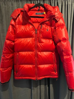 Röd pufferjacka från Polo Ralph Lauren - Snygg röd pufferjacka från Polo Ralph Lauren med huva och två sidofickor med dragkedja. Jackan har en mörkblå logga på bröstet och quiltad design. Perfekt för kalla dagar och har en modern look med glansigt material.