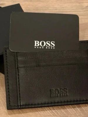 Svart plånbok från Hugo Boss - Snygg och stilren svart plånbok från Hugo Boss i äkta skinn. Plånboken har flera fack för kort och är prydd med diskret Boss-logga framtill. Perfekt för dig som gillar minimalistisk design och vill ha koll på dina kort.