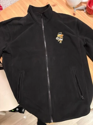 Svart fleecejacka AIK med dragkedja - Svart fleecejacka med hel dragkedja och två fickor med dragkedja. Den är helt ny aldrig använd ny pris är 550 och den går endast att köpa på norra stå ifrån sol invictus . Säljes pga köpte för liten storlek 