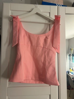 Rosa linnetopp med breda axelband  - Säljer en ljusrosa linnetopp med breda axelband som har en snygg detalj med knyt. Toppen har en rak och något figurnära passform, ärmlös design och är tillverkad i ett slätt bomullsmaterial. Perfekt för sommarens alla tillfällen! Dragkedja på sidan och stor i storlek 