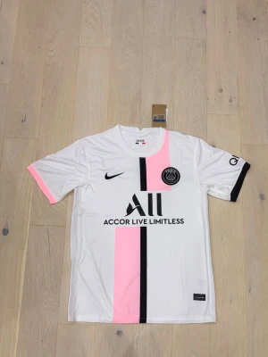 PSG football jersey - Bilderna visar en vit och rosa Paris Saint-Germain fotbollströja från Nike, inte några skor. Tröjan har svarta detaljer och klubbens emblem på bröstet.