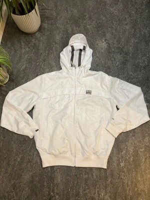 Vit vindjacka från McKenzie NYC - Snygg vit vindjacka från McKenzie NYC med huva och dragkedja framtill. Jackan har diskret mönster med NYC-tryck, ribbade muddar och meshfoder på insidan. Perfekt för dig som gillar streetwear och vill ha en lätt jacka med coola detaljer.