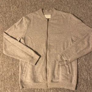 Grå ziptröja från Bread & Boxers - Snygg grå ziptröja från Bread & Boxers i storlek S. Tröjan har lång ärm, ribbade muddar och en hel dragkedja framtill. Tillverkad i mjuk bomull som är skön att ha på sig. Perfekt för chill dagar eller när du vill ha en enkel och clean look.