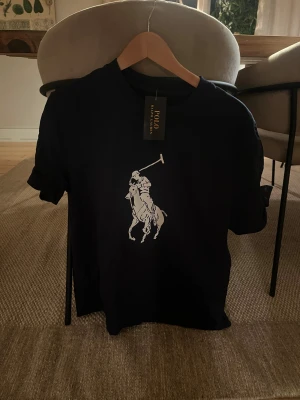 mörkblå t-shirt från Polo Ralph Lauren - Mörkblå t-shirt från Polo Ralph Lauren med klassisk vit polospelare-logga på bröstet. T-shirten har rund hals, korta ärmar och är gjord i mjuk bomull. Perfekt för en clean och stilren look.