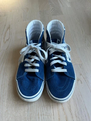 Blå Vans Sk8-Hi sneakers strl 36 - Säljer ett par blå Vans Sk8-Hi sneakers i storlek 36 med vit snörning och checkerboard-mönster med Vans-logga på sidan. Skorna har mockadetaljer och klassisk vit sula med blå rand. Höga och riktigt snygga för dig som gillar streetstyle.