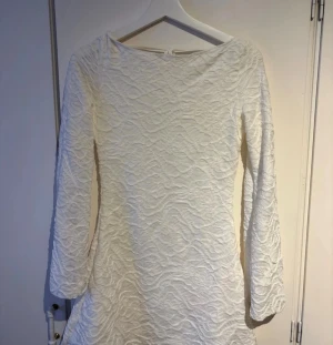 Vit kort klänning  - Säljer en vit kort klänning med långärmade ärmar. Klänningen har en öppen rygg med knytband upptill och en lätt utsvängd form. Köpt i år och använts en gång! Storlek S