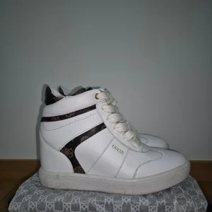 Guess vita sneakers  - Säljer ett par vita sneakers från Guess med högt skaft och snörning. Skorna har svarta och bruna detaljer med logomönster på skaftet och sidan. Tillverkade i skinnimitation med platt sula och rund tå. Snygga och trendiga för dig som gillar streetstyle.