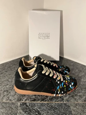 Svarta Maison Margiela paint splatter skor - Maison Margiela sneakers i svart skinn och mocka med beige insida och snörning. Skorna har en unik färgstänksdesign i blått, gult, rött och vitt över hela ovandelen. Sulan är gummibrun med Maison Margiela-logga. Riktigt coola och konstnärliga sneakers för dig som vill sticka ut.