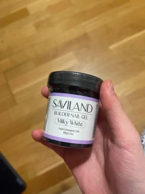 Saviland Builder Nail Gel Milky White - Säljer en burk Saviland Builder Nail Gel i färgen Milky White. Gelen är perfekt för att bygga och förlänga naglar, ger en mjuk och mjölkvit finish som syns på bilden. Kommer i en rund plastburk med svart lock, innehåller 100g.