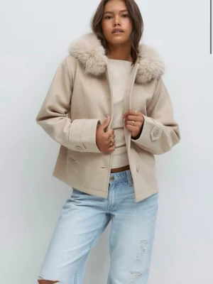 Beige kappa med pälsdetalj - Snygg beige kappa med fluffig pälsdetalj på kragen. Jackan har en rak passform, knappar framtill och breda muddar med knappdetaljer. Perfekt för dig som vill ha en stilren och trendig look under kyliga dagar.