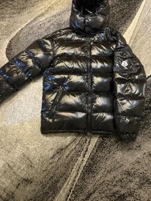 Svart Moncler pufferjacka med huva - Säljer en svart pufferjacka från Moncler med glansig finish och huva. Jackan har dragkedja framtill, två sidofickor med dragkedja och Moncler-logga på ärmen. Insidan är grå och jackan är vadderad för extra värme. Perfekt för kalla dagar.