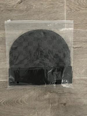 Rutig grå Louis Vuitton mössa - Snygg mössa från Louis Vuitton i grått med rutigt mönster och svart uppvikt kant. LV-logga framtill och diskret tagg på insidan. Materialet är mjukt och känns lyxigt, perfekt för dig som vill ha något stilrent och exklusivt.