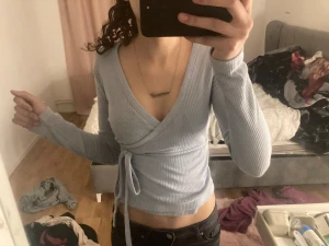 Ljusblå omlottopp med knyt - Säljer en ljusblå omlottopp med ribbad struktur och långa ärmar. Toppen har v-ringning och knyt i midjan som ger en snygg passform. Perfekt att styla med jeans eller kjol för en chill look.