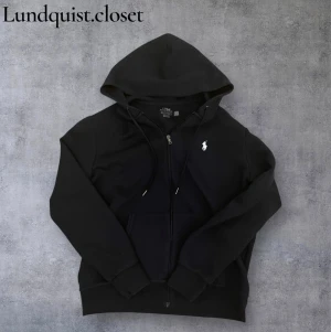 Mörkblå hoodie med dragkedja från Polo Ralph Lauren - Mörkblå hoodie från Polo Ralph Lauren med klassisk vit logga på bröstet. Tröjan har huva med snörning, dragkedja framtill och en stor ficka på magen. Storlek L men passar också bra som storlek M