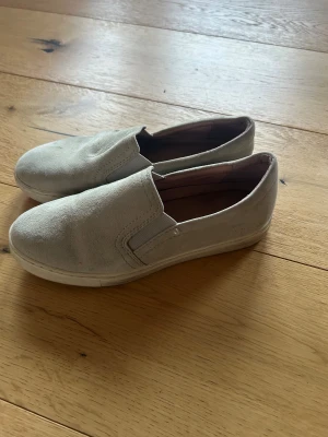 Beige slip-on sneakers från Dash - Säljer ett par beige slip-on sneakers från Dash med ovandel i mocka och elastiska paneler på sidorna för enkel på- och avtagning. Skorna har platt sula och rund tå, vilket gör dem riktigt sköna att glida runt i. Perfekt för dig som gillar enkel och clean stil. Nypris ca 900: 