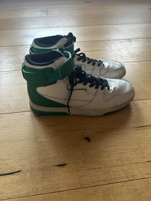 Grönvita high-top sneakers från Zara - Säljer ett par grönvita high-top sneakers från Zara med svart snörning och perforerad tå. Skorna har gröna detaljer och en kraftig platt sula med grönt mönster undertill. Materialet är syntet och skinnliknande, med vadderad kant och kardborreband vid ankeln.