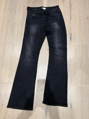 Svarta bootcut jeans i stretch från Gina Young  - Svarta jeans från Gina Young i storlek 152 med bootcut-ben och klassisk femficksdesign. Jeansen har snygga slitningar framtill och bakfickor med knappdetaljer. Mycket mjukt och stretchigt jeanstyg.