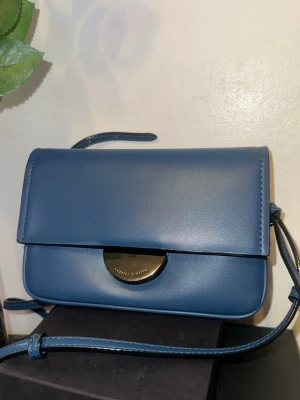 Blå axelväska från Charles & Keith - Snygg blå axelväska från Charles & Keith med stilren design och silverfärgad metallknäppning framtill. Väskan har slät yta i skinn och justerbar axelrem. Perfekt storlek för det viktigaste och passar till många olika outfits.