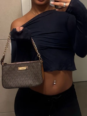 Brun handväska från Michael Kors - Säljer en snygg brun handväska från Michael Kors med guldiga kedjedetaljer och dragkedja. Väskan har MK-logga i mönstret och en metallplatta framtill. Perfekt storlek för det viktigaste och har en lyxig vibe med sitt guldiga axelband.