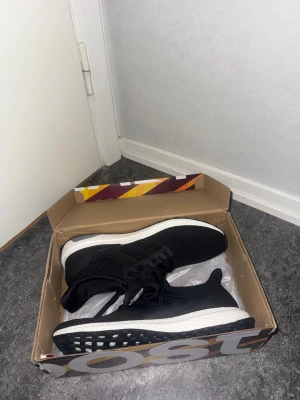 Adidas Ultraboost svarta sneakers - Svarta Adidas Ultraboost sneakers med vit Boost-sula och stickad ovandel. Skorna har snörning och en platt sula med rund tå. Perfekta för dig som gillar stilrena och bekväma skor med sportig vibe. Säljes i originalkartong.