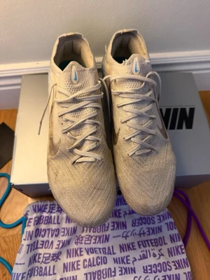 Nike Mercurial fotbollsskor elite - Nike Mercurial fotbollsskor i ljusbeige med silvriga swoosh-loggor och snörning. Ovandelen är stickad och sulan har en metallicblå och lila finish med dobbar för gräsplan. Skorna har rund tå och platt sula, samt detaljer i blått och vitt på hälen.