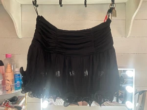 Svart volangkjol med dragkedja - Säljer en svart kortkjol med volange och snyggt veckad struktur. Kjolen har en dragkedja i sidan och är gjord i ett lätt, transparent tyg som ger en cool och luftig känsla. Perfekt för dig som gillar detaljer och vill sticka ut med en unik look.