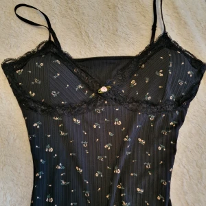 Söt blommig linnetopp med spets 3 st - 3 st rosa,svart,vit Supersöta linnetoppar med smala axelband och spetskant vid bysten. Finns i svart, rosa och vit med små blommor och en liten rosett framtill. Ribbad struktur i tyget och mjuk, stretchig känsla. Perfekt för att ge din outfit en gullig och romantisk vibe.