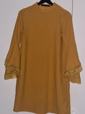 Ockerfärgad klänning från Dilvin  Woman - Snygg ockerfärgad klänning från Dilvin Woman med långa trumpetärmar och spetsdetaljer vid ärmsluten. Rund halsringning med dekorativa knappar på axeln. Klänningen har en rak passform och är tillverkad i ett mjukt tyg som känns skönt mot huden.