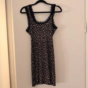 Blommig klänning  - Svart ribbad klänning från H&M Divided i storlek S med små rosa och ljusgröna blommor. Klänningen är ärmlös och har spetskant runt halsringningen och axelbanden. Mjuk och stretchig bomullskvalitet, perfekt för dig som gillar söta detaljer och blommigt.
