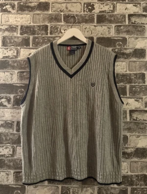 Grå stickad väst från Chaps Ralph Lauren - Grå stickad väst från Chaps Ralph Lauren med ribbad struktur och mörkblå kant vid v-ringad hals och ärmöppningar. Västen har ett broderat emblem på bröstet och är tillverkad i 100% bomull. Perfekt för lager-på-lager och snygg till skjorta.