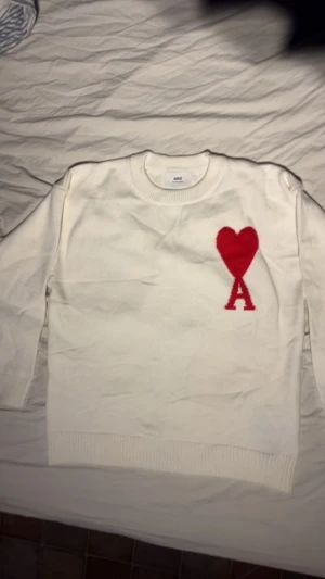 Vit stickad tröja från Ami Paris - Vit stickad tröja från Ami Paris med ett stort rött hjärta och bokstaven A broderat på bröstet. Tröjan har rund hals, ribbade muddar och är långärmad. Perfekt för dig som gillar stilrena plagg med en unik detalj.