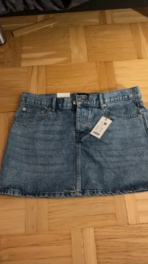 Blå jeanskjol från 157, M - Säljer en klassisk blå jeanskjol från 157 i modell Maddy. Kjolen har fem fickor, bälteshällor och knapp- samt dragkedja framtill. Den är kort och har en rak passform som ger en cool och avslappnad vibe. Perfekt att styla med sneakers eller boots.