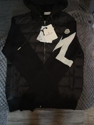 Svart pufferjacka Moncler med huva - Svart pufferjacka från Moncler med huva och snörning. Jackan har dragkedja framtill och ett coolt vitt zigzag-mönster på ena ärmen. Märkespatch på ärmen och ribbade muddar. Materialet är polyester och dun, perfekt för kyliga dagar. Passar m 