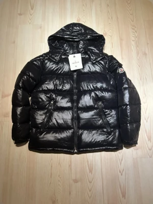 Moncler Maya  - Säljer en svart pufferjacka från Moncler med glansig finish och huva. Jackan har dragkedja framtill, två snedställda fickor med dragkedja och Moncler-logga på ärmen. Insidan har ett coolt serietryck. Perfekt för dig som gillar streetstyle.