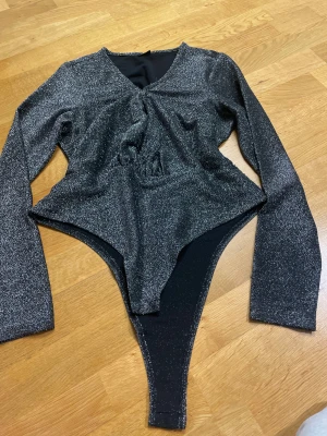 Glittrig svart body från Gina Tricot - Säljer en svart glittrig body från Gina Tricot i storlek XL. Bodyn har lång ärm, v-ringad hals och knäppning med tre svarta knappar i grenen. Materialet är syntet med glittereffekt som ger en snygg look. Perfekt för att sticka ut med lite extra bling.