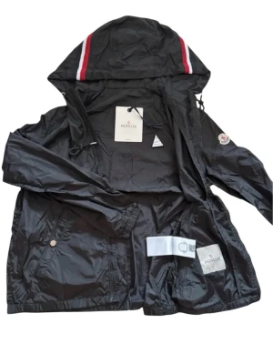 Svart vindjacka från Moncler med huva - Svart vindjacka från Moncler med huva och snygga röda och vita ränder på toppen. Jackan har dragkedja, snörning i huvan och Moncler-logga på ärmen. Materialet är lätt och syntetiskt, perfekt för blåsiga dagar. Fickor med tryckknappar och stilren design.