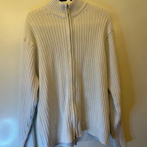 Vit ribbad kofta från Kitaro  - Säljer en vit ribbstickad kofta från Kitaro i storlek XL. Tröjan har hel dragkedja framtill och en ståkrage med marinblå insida. Materialet är stickad bomull och modellen är normal passform med långa ärmar.
