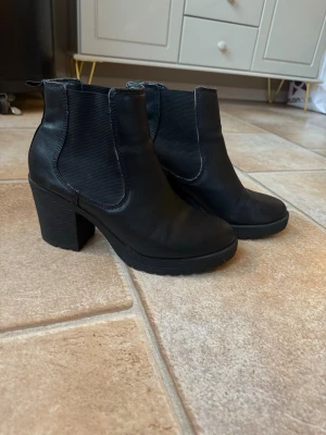 Svarta chelsea boots med klack storlek 39  - Säljer ett par svarta chelsea boots med grov sula och hög klack. Skorna har rund tå, elastiska paneler på sidorna och är tillverkade i syntetmaterial med skinnlook. Perfekt för dig som gillar chunky boots med lite höjd. Ca 8 cm klack. Sparsamt använda. Finns lite färg slitage se sista bilderna! Nypris 699:- 