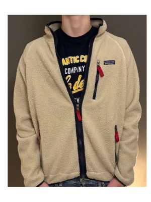 Beige fleecejacka från Patagonia - Säljer en beige fleecejacka från Patagonia med mörkblå detaljer och röda dragkedjor. Jackan har hög krage, tre fickor med dragkedja och en loose passform. Perfekt för dig som gillar chill och comfy stil.