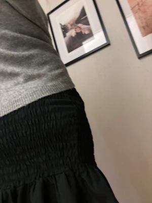 Svart kortkjol med volang - Säljer en svart kortkjol med smockad midja och volangkant nedtill. Kjolen har en tight passform upptill och är gjord i ett lätt, mjukt tyg som ger en snygg siluett. Perfekt för dig som gillar stilrena plagg med lite extra detalj. Fråga gärna om mått!