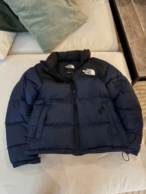 Blå pufferjacka från The North Face - Säljer en mörkblå pufferjacka från The North Face med svart axelparti och logga på både fram- och baksidan. Jackan har dragkedja, två sidofickor och elastiska muddar vid ärmslut. Perfekt för kalla dagar och riktigt snygg till streetwear.