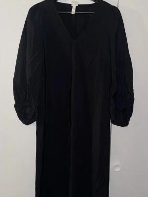 Svart långklänning med v-ringning - Svart långklänning från H&M med v-ringad hals och långa puffärmar. Klänningen har en enkel, rak siluett och är tillverkad i ett lätt, skrynkligt syntetmaterial som ger en cool och avslappnad vibe. Perfekt för dig som gillar minimalistisk stil.