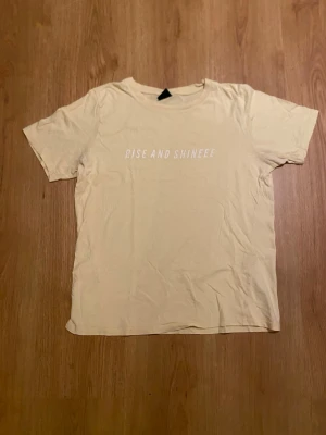 Gul t-shirt med text - Säljer en gul t-shirt med trycket 'RISE AND SHINEEE' i vitt på bröstet. T-shirten har en klassisk rund hals och korta ärmar. Materialet känns mjukt och lätt, perfekt för chill dagar. Enkel och stilren design som funkar till jeans eller shorts.
