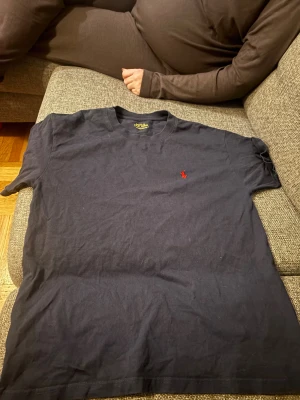 Svart t-shirt Polo Ralph Lauren - Svart t-shirt från Polo Ralph Lauren med klassisk röd broderad logga på bröstet. Modellen är kortärmad och har en rund hals. Materialet är mjuk bomull och passformen är slim. Perfekt för dig som gillar stilrena och ikoniska plagg.