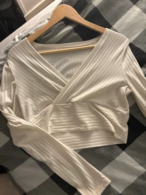 Vit omlott croppad topp från SHEIN - Säljer en vit croppad omlottopp från SHEIN i storlek M. Toppen har långa ärmar, ribbad struktur och en snygg omlottdesign med v-ringad hals. Perfekt för dig som gillar stilrena och trendiga plagg.