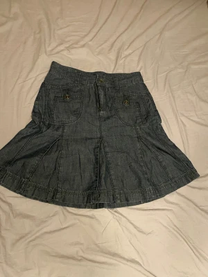 Mörkblå jeanskjol med fickor och dragkedja - Säljer en mörkblå jeanskjol i kort modell med flera fickor fram och bak, samt snygga detaljer som knappar och dragkedja. Kjolen har bälteshällor och en lätt utsvängd form som ger en cool och avslappnad vibe.