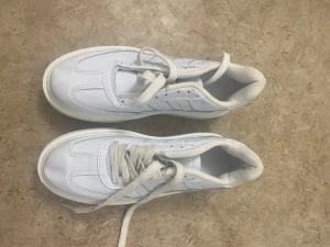 Vita sneakers från Adidas, strl 39 - Fräscha vita sneakers från Adidas med klassiska ränder på sidan och rund tå. Skorna har snörning och en platt sula i vitt. Tillverkade i syntetmaterial med textilfoder för extra komfort. Perfekt för dig som gillar stilrena och sportiga skor.